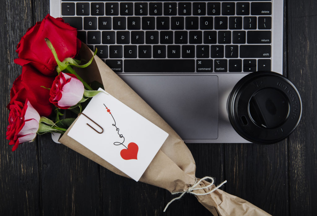 vue de dessus d'un bouquet de roses rouges en papier craft avec une carte postale attachée, posé sur un ordinateur portable avec une tasse de café en papier sur fond de bois foncé