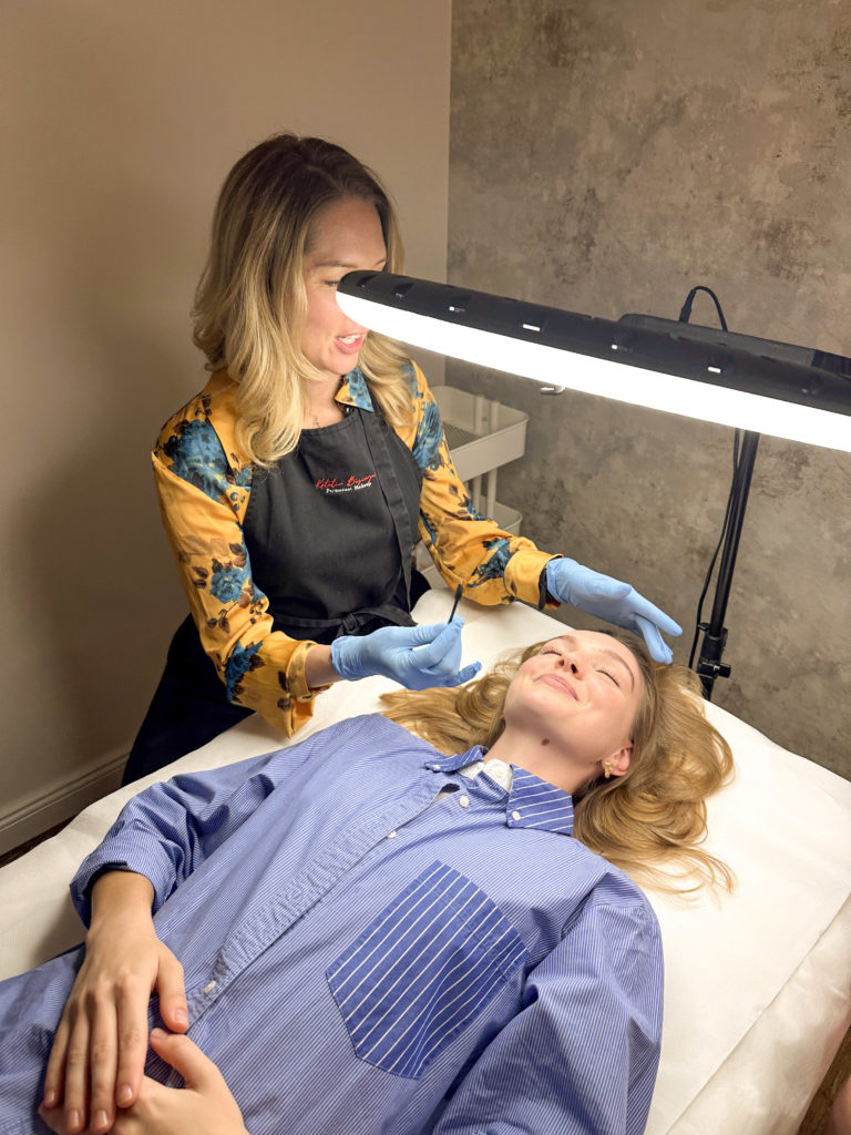 A beauty technician in gloves carefully performs a facial procedure on a client under bright ring lights. The setting is calm and professional.