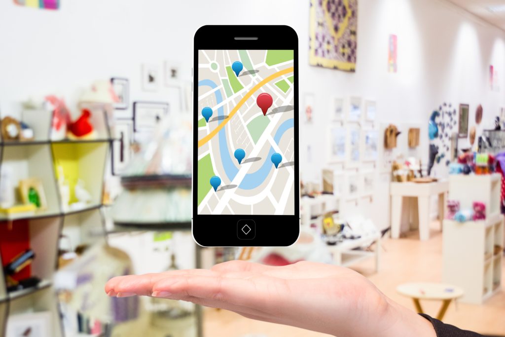 A hand holds a smartphone displaying a map with colored pins, illustrating centralized scheduling for multiple locations. In the background, a brightly lit shop with various items.