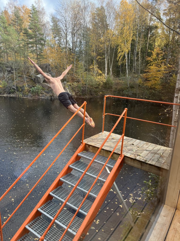 Eine Person in Badehose springt von einer Metalltreppe mit orangefarbenem Geländer in einen ruhigen See.  