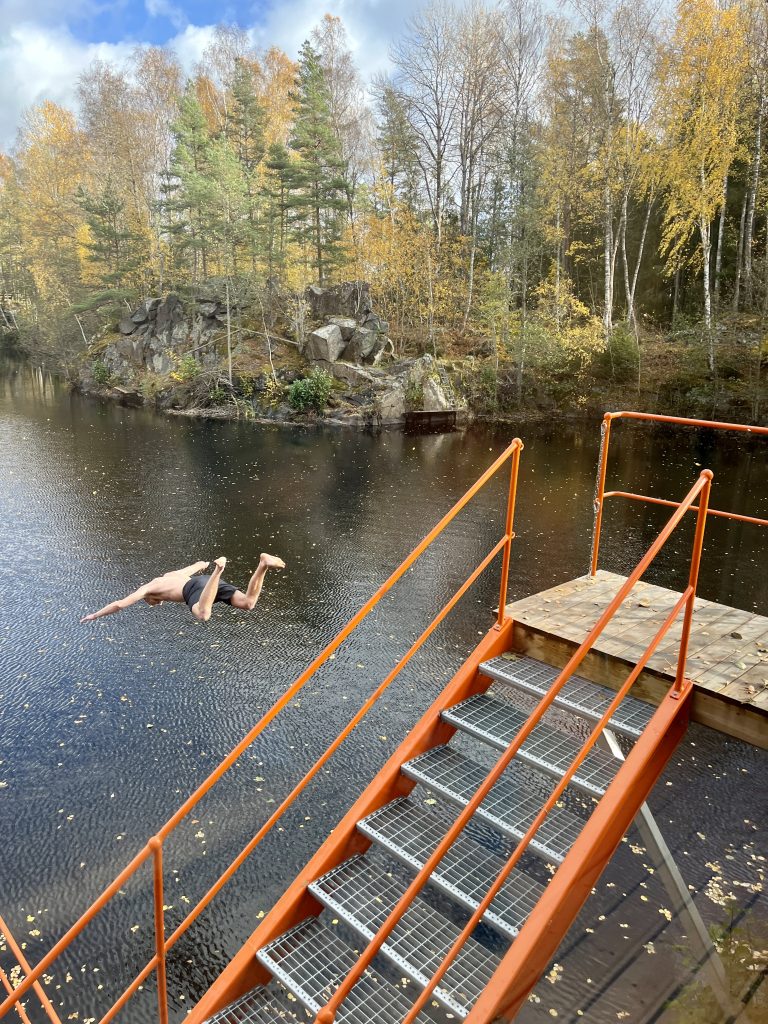 Eine Person stürzt sich aus der Luft in einen ruhigen See, der von Herbstbäumen umgeben ist.  
