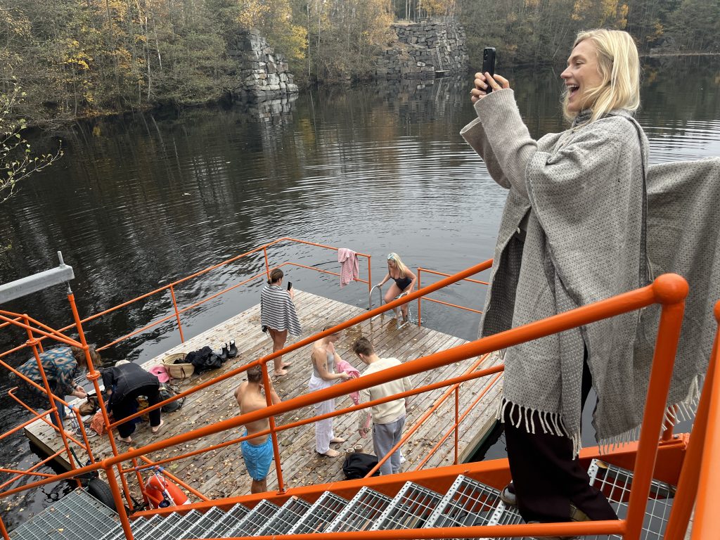 Eine lächelnde, in eine Decke gehüllte Frau fotografiert eine Gruppe von Menschen auf einem orangefarbenen Steg an einem ruhigen See.  