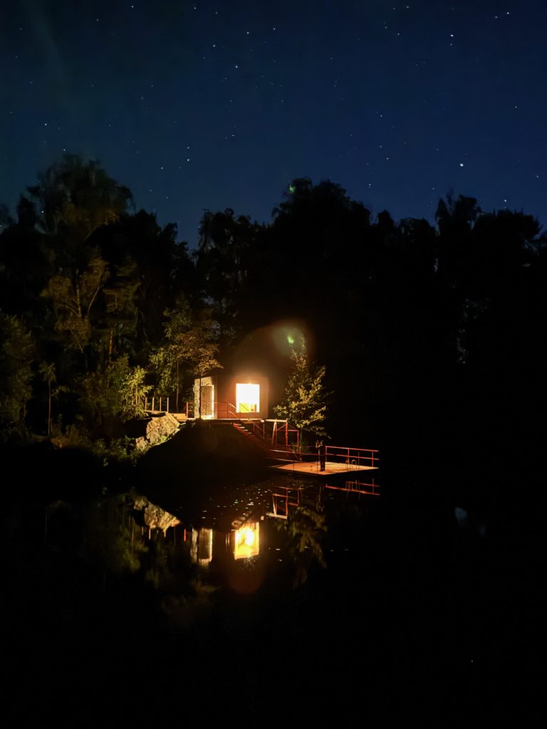 Ein warm beleuchtetes Gebäude steht an einem bewaldeten Seeufer, dessen Spiegelbild im ruhigen Wasser schimmert. Der dunkle Himmel darüber ist mit Sternen übersät. 