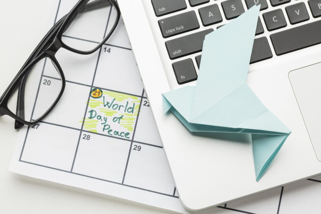 Calendar with "World Day of Peace" on the 21st, glasses, and a blue origami bird on a laptop. The scene evokes calm and contemplation.