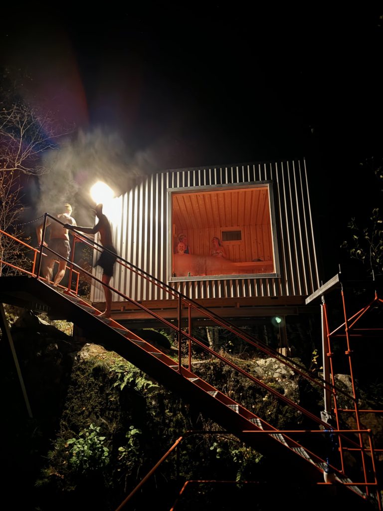 Die Gemeinschaftssauna Gylsboda Kallbadhus leuchtet in einem würfelförmigen Gebäude bei Nacht, zwei Personen auf einer roten Treppe.