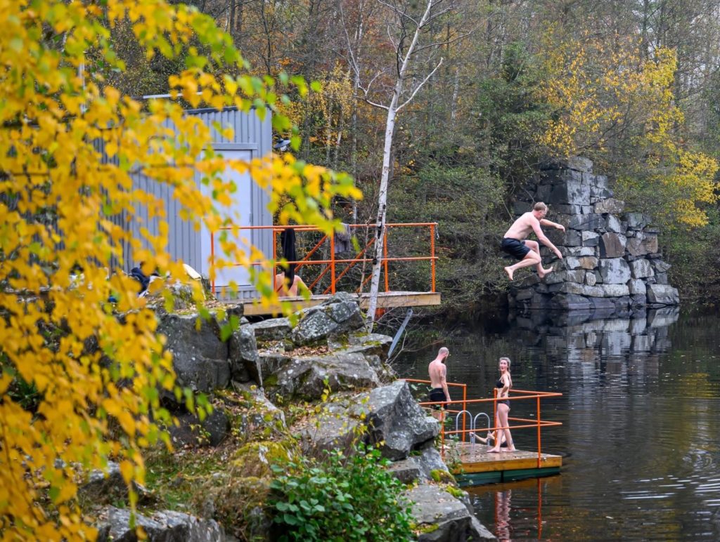 Eine Person springt in einen See im Gylsboda Kallbadhus, umgeben von herbstlichen Bäumen und felsigen Klippen, während andere das gemeinsame Saunaerlebnis in Gylsboda genießen