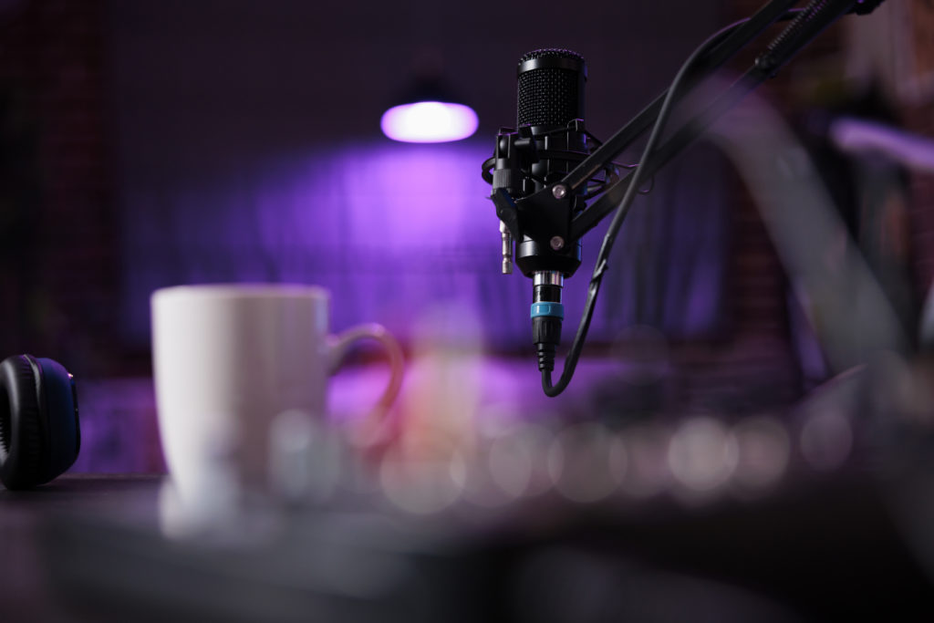 Podcast studio with a black microphone, headphones, and white mug on a table, lit with soft purple lighting, illustrating online scheduling for music studios.
