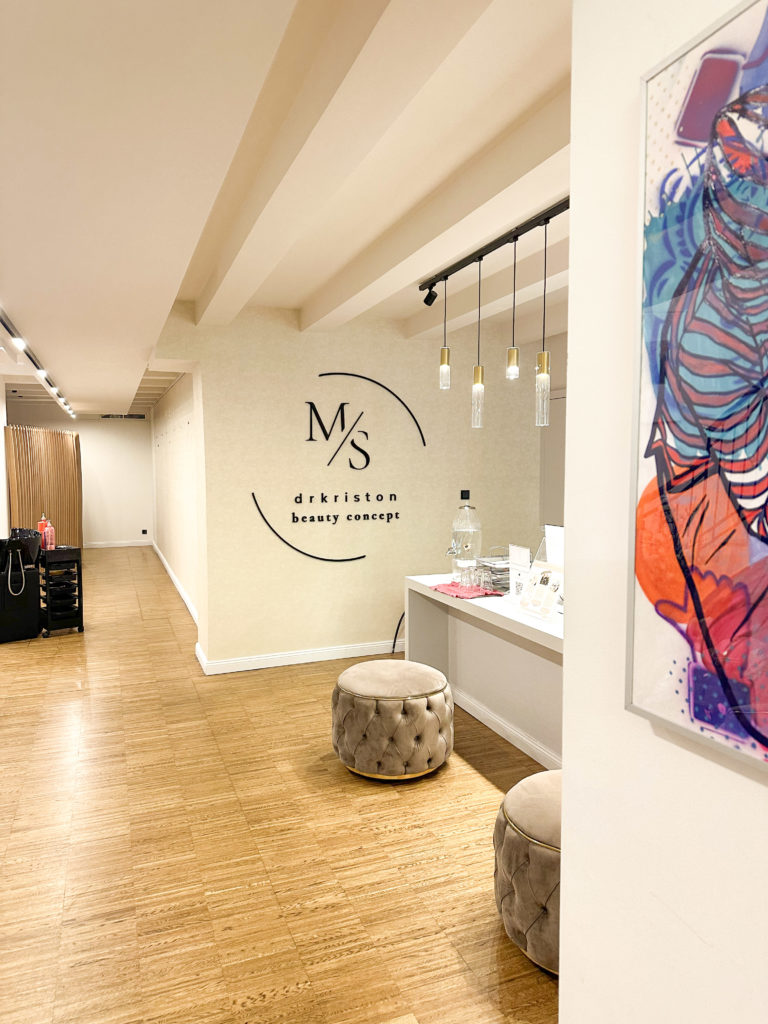 Modern beauty salon interior with wooden floors, plush stools, and minimalist decor. Soft lighting and a hallway with branded "M/S dr kriston beauty concept" logo.
