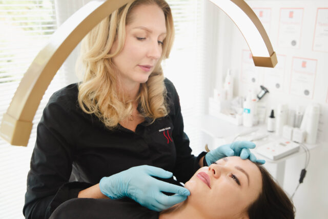 A beautician with blonde hair and wearing blue gloves carefully examines a woman's face under a curved light in a clinical, serene setting.
